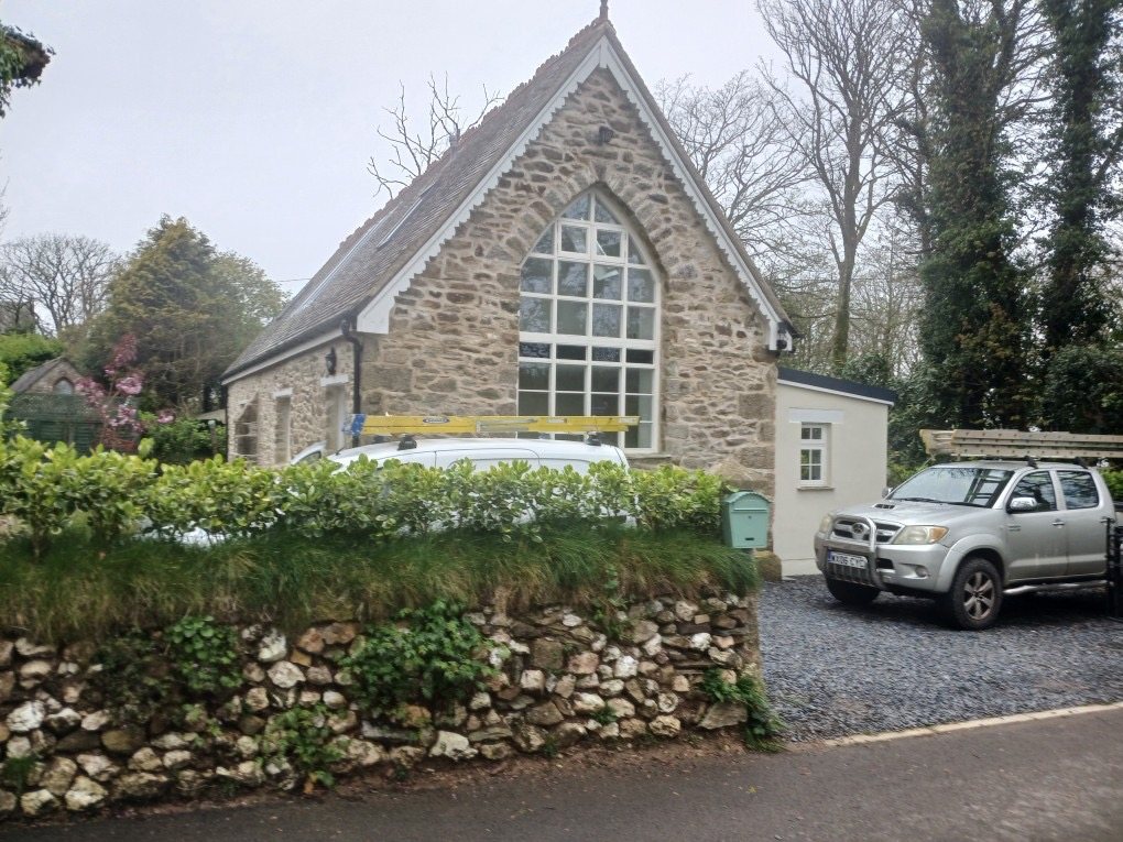 Restored stone building with a tall arched window in the gable, behind a stone hedge